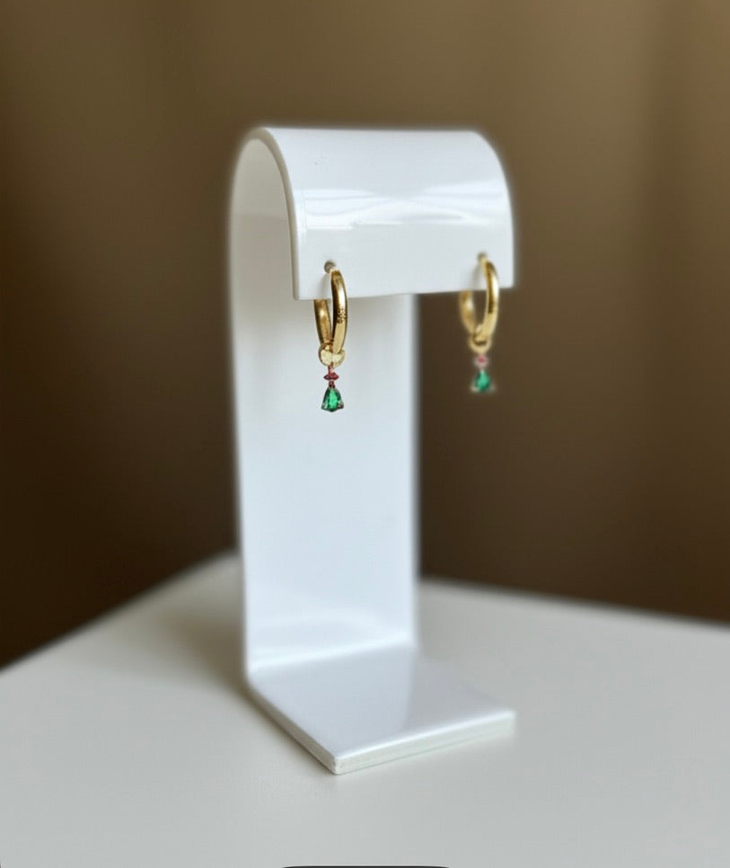 Gold hoop earrings with green gemstones on a white stand against a brown background